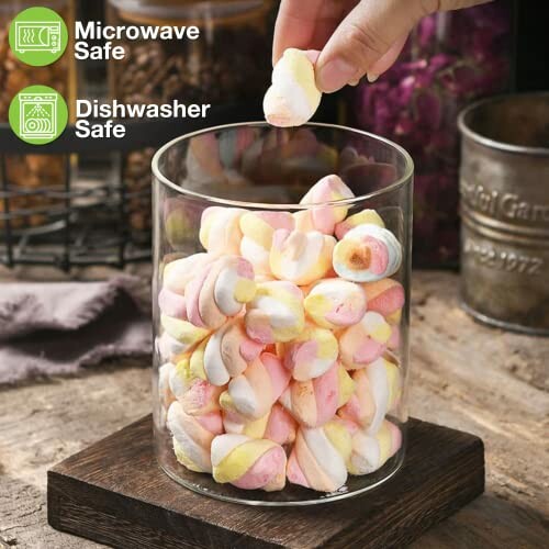 Hand reaching for colorful marshmallows in a glass jar on a wooden surface, with microwave and dishwasher safe icons.